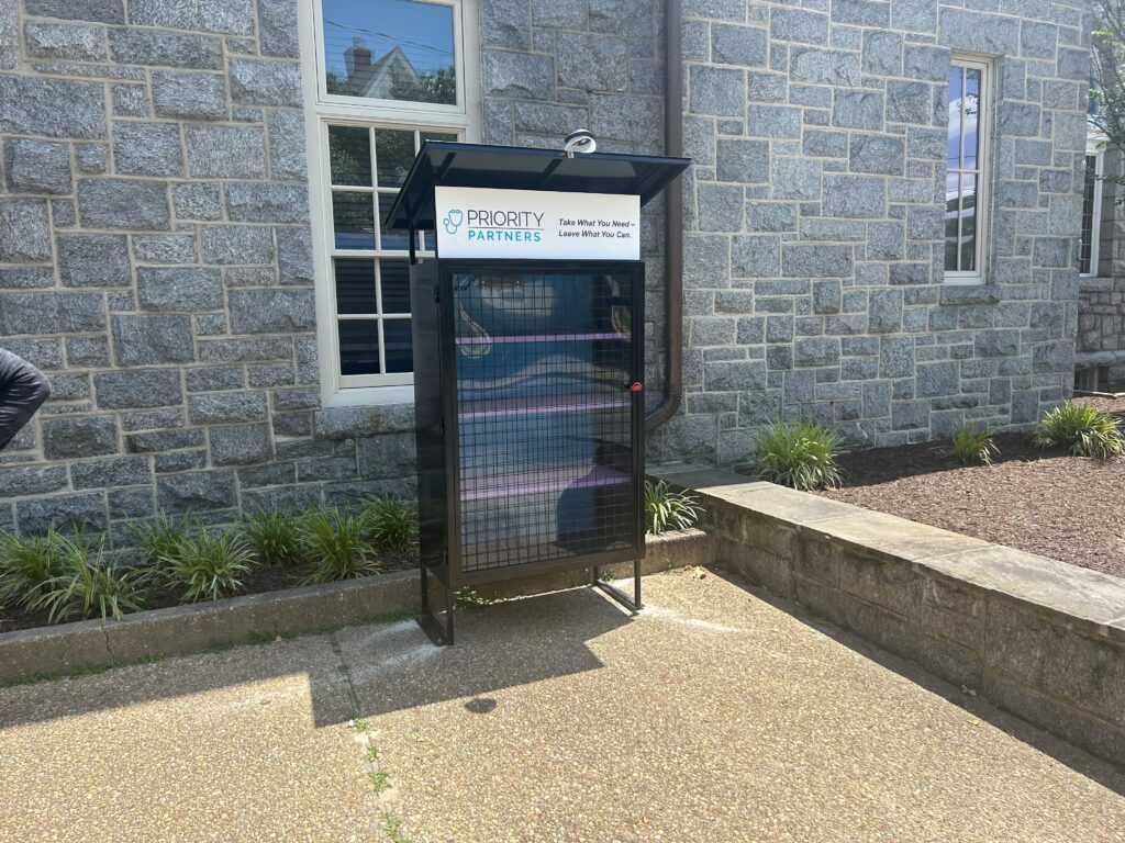 Recently built empty pantry cupboard featuring an Priority Partners' logo on its side panel, set against a stone building and greenery.