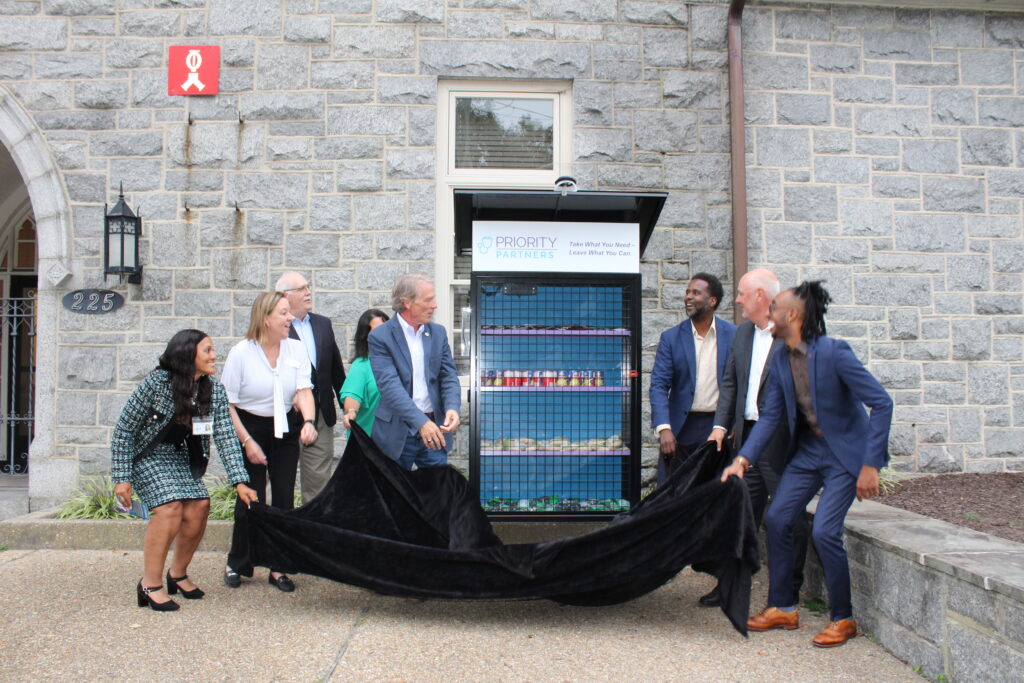 Group of people unveiling a food pantry display from Priority Partners in an event outside a stone building.