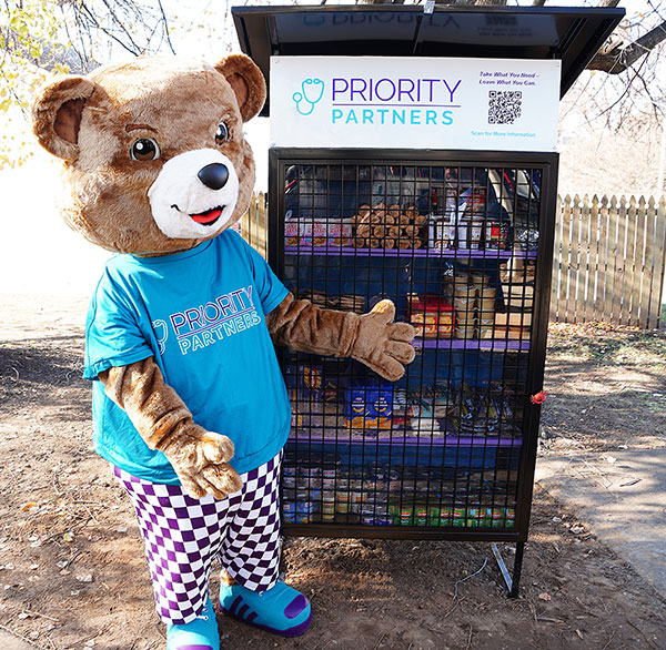 Cubby the bear mascot for Priority Partners standing next to a mini food pantry